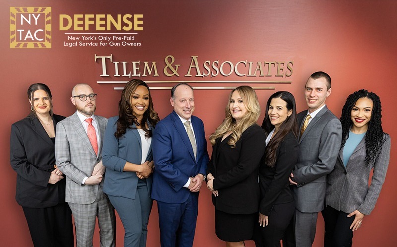 A group portrait of eight legal professionals from Tilem & Associates standing together in business attire against a reddish-tan wall with gold lettering reading "TILEM & ASSOCIATES". In the top-left corner, logos for "NY TAC" and "DEFENSE" read "New York's Only Pre-Paid Legal Service for Gun Owners."